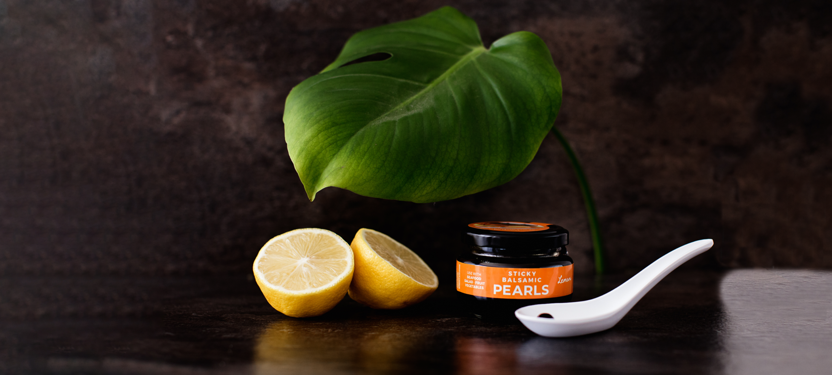 A jar of Sticky Balsamic Lemon Pearls with two half lemons, and a white spoon with a single Sticky Balsamic Pearl, with a leaf and dark background. Balsamic Vinegar