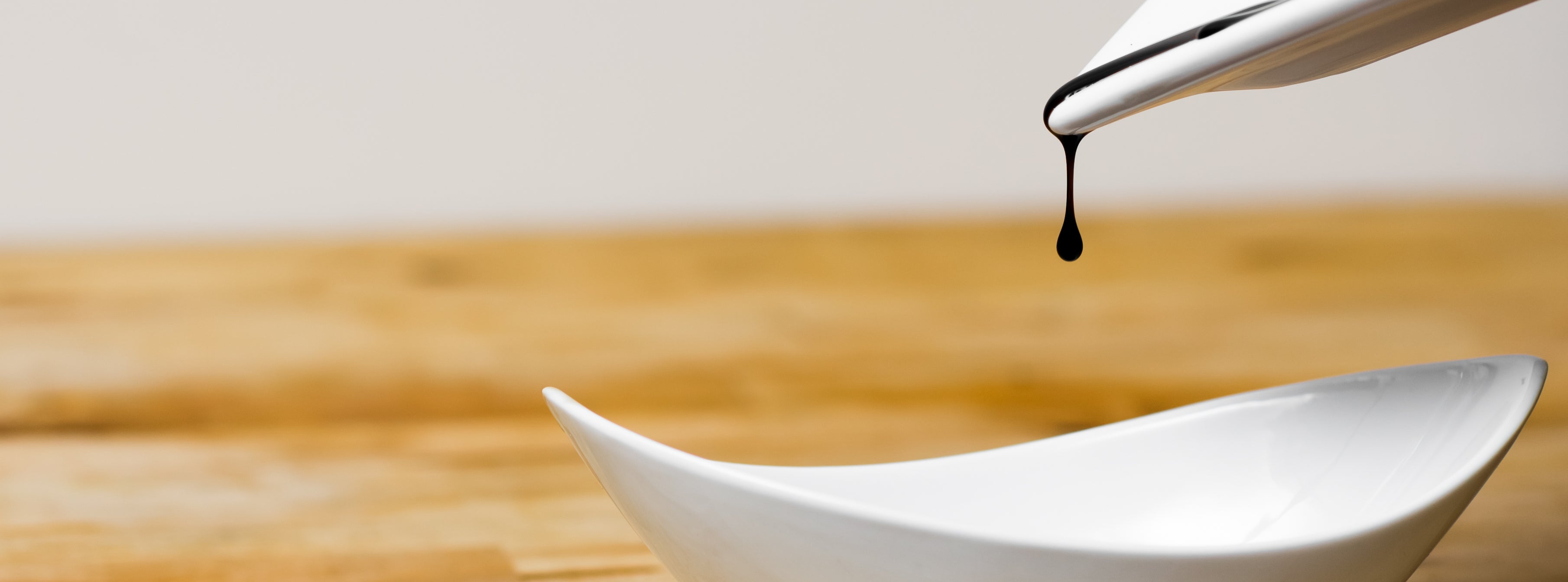 A single drip of Sticky Balsamic which looks artistic, being slowly poured from one white vessel to another white vessel, with a white and wooden background.
